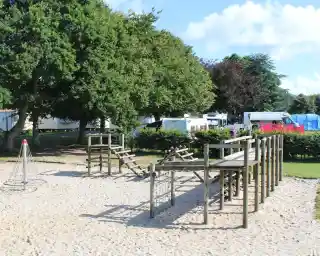 A children's playground featuring wooden climbing structures, a rope climbing frame, and a sandy area, surrounded by trees and camping vehicles in the background.