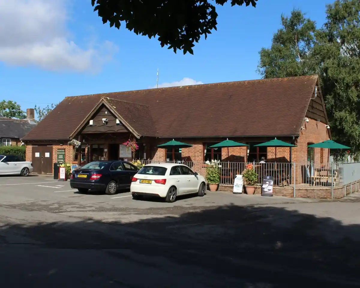 A brick building with a sloped roof, featuring outdoor seating under green umbrellas. Two cars are parked in front, and there are planters with flowers visible. Trees and a blue sky with clouds are in the background.