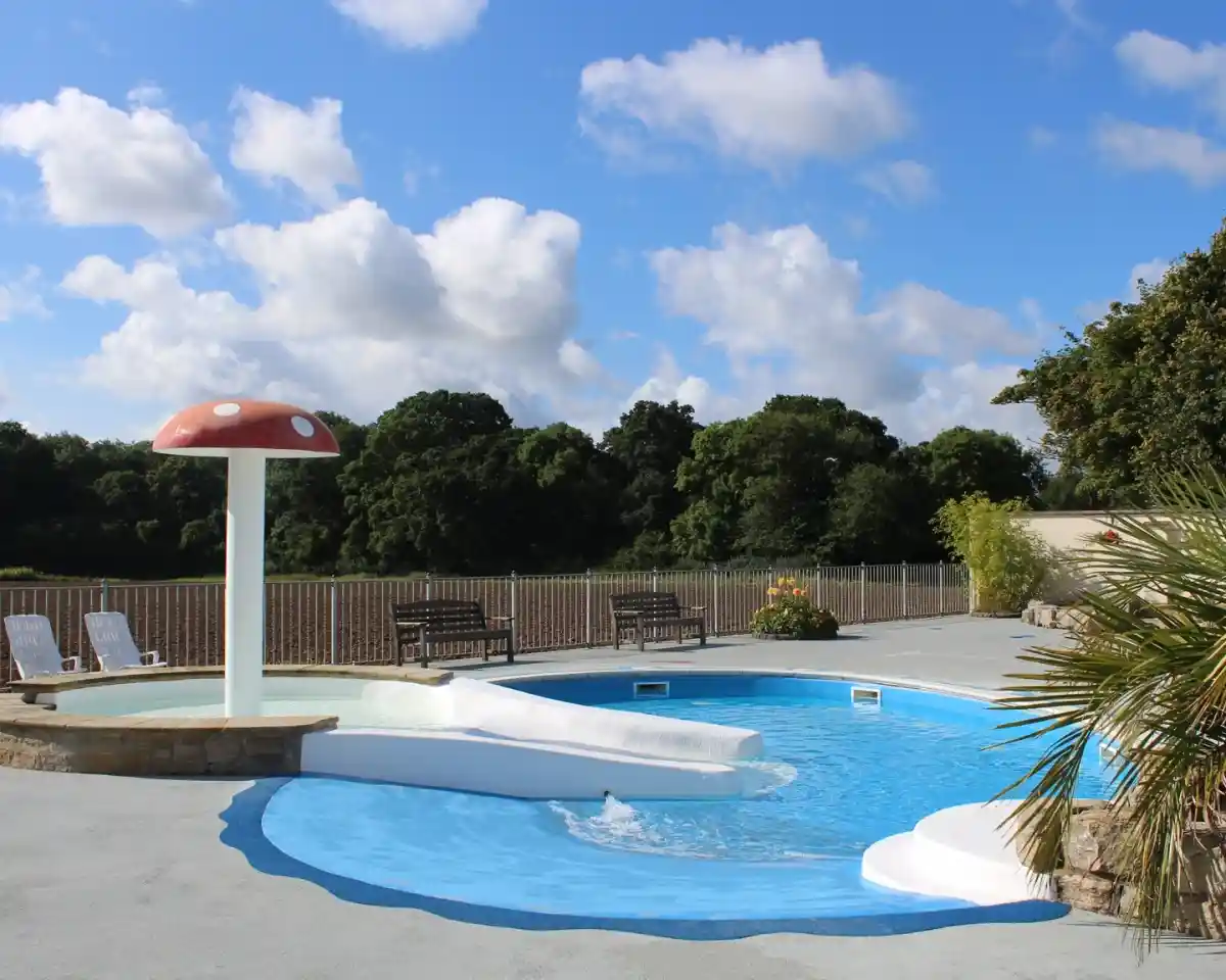 A bright, inviting pool area features a mushroom-shaped water slide, with a shallow section and a gentle waterfall. Lush trees line the background under a partly cloudy sky. Benches are positioned nearby, and tropical plants add a touch of greenery.