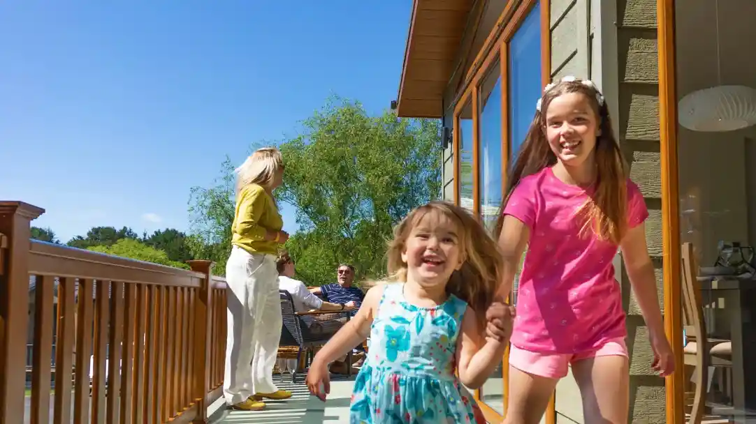 Two young girls hold hands and run joyfully on a sunny deck. One girl wears a blue dress while the other is in a pink outfit. In the background, adults are seated and engaged in conversation. Bright blue sky and greenery enhance the cheerful atmosphere.