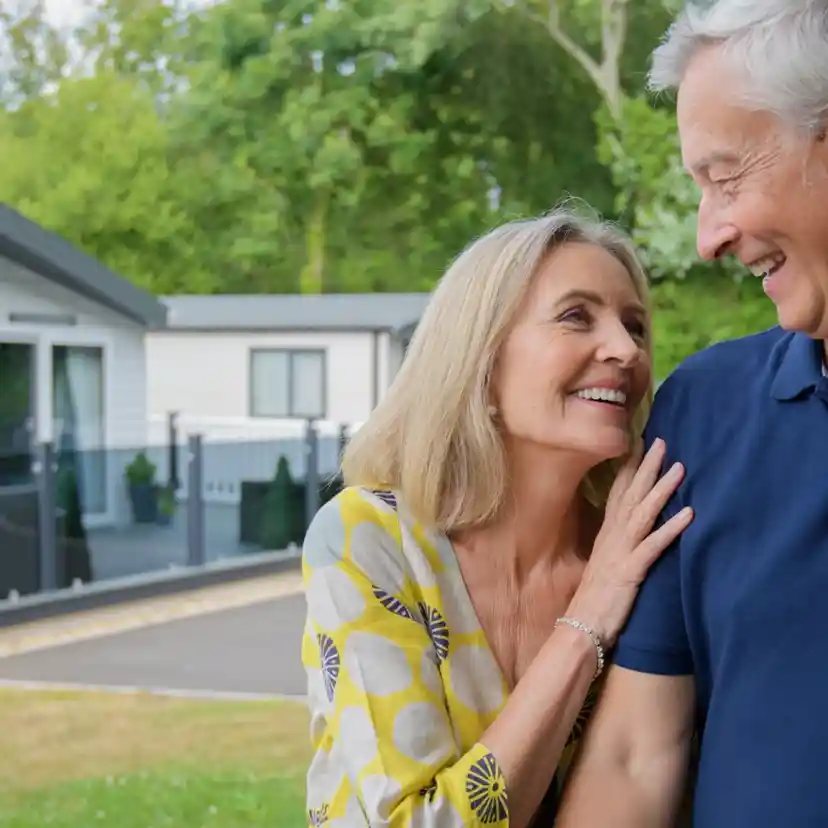 A happy older couple smiles at each other outdoors, with a modern house and lush greenery in the background. The woman leans affectionately on the man's shoulder.