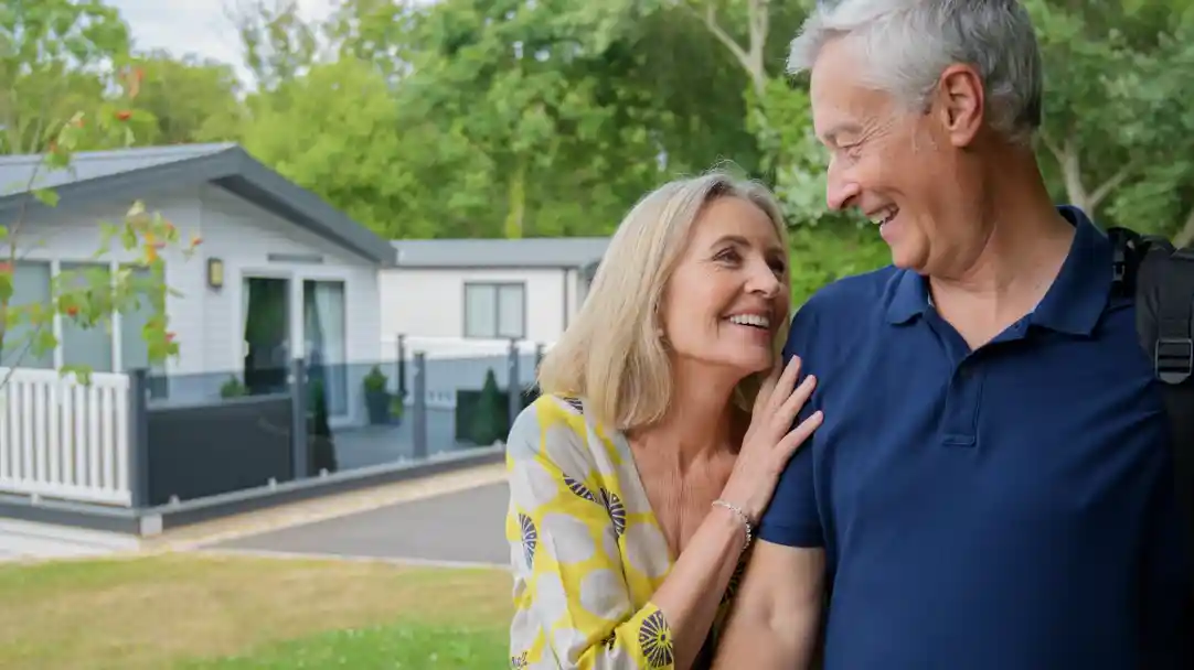 A happy older couple smiles at each other outdoors, with a modern house and lush greenery in the background. The woman leans affectionately on the man's shoulder.