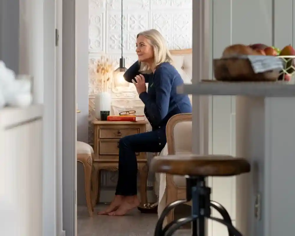 A woman with blonde hair sits at a small desk in a cozy, well-lit room. She has a relaxed expression and is looking to the side, with a lamp illuminating her workspace. A wooden stool and some kitchen items are visible in the foreground.