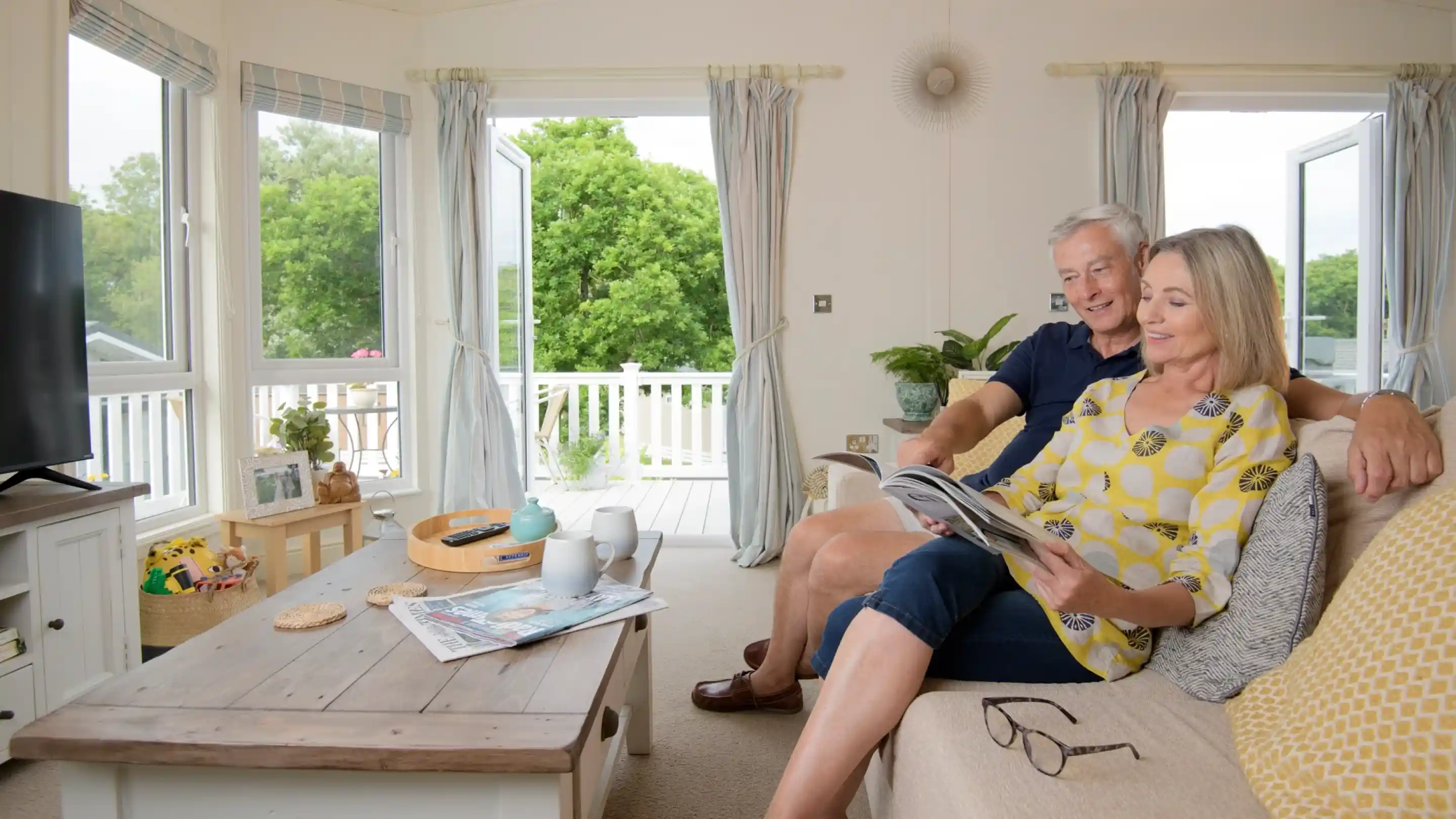 A couple sits together on a couch in a bright, cozy living room. The woman reads a magazine while the man smiles at her. Sunlight filters in through open windows, revealing greenery outside. A coffee table in front holds a cup, a magazine, and a small decorative item.