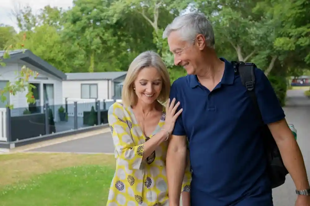 A smiling couple walks along a path in a scenic area, enjoying each other's company. The woman, wearing a yellow patterned dress, lightly touches the man's arm as they share a moment of joy. Trees and modern cabins can be seen in the background.
