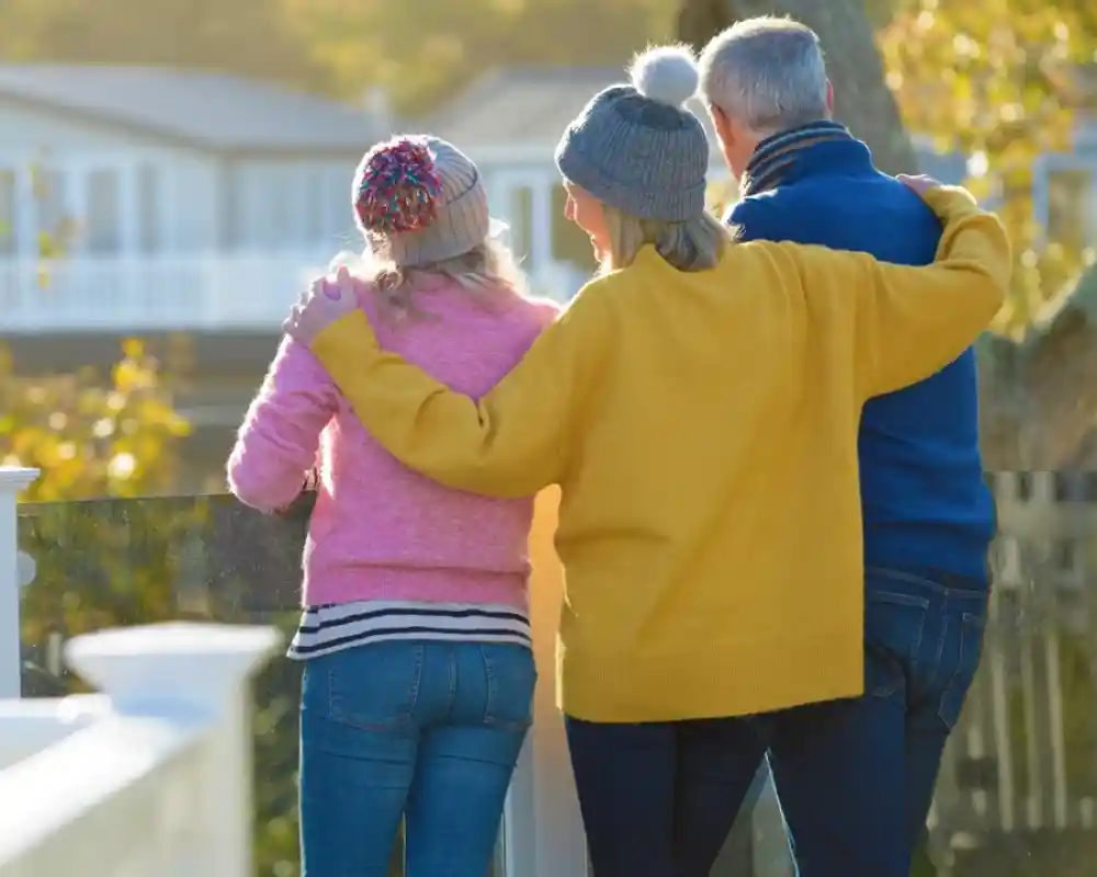 Three people stand side by side, overlooking a scenic view. Two women wear colorful sweaters—one pink and one yellow—while a man in a blue jacket stands beside them. They have their arms around each other, enjoying a sunny day surrounded by trees.