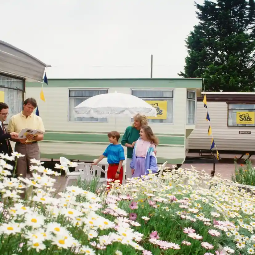 A family stands in a flower-filled garden in front of two mobile homes. One adult reads a brochure to another adult while two children listen. A girl holds an umbrella, and colorful flags adorn the mobile homes.