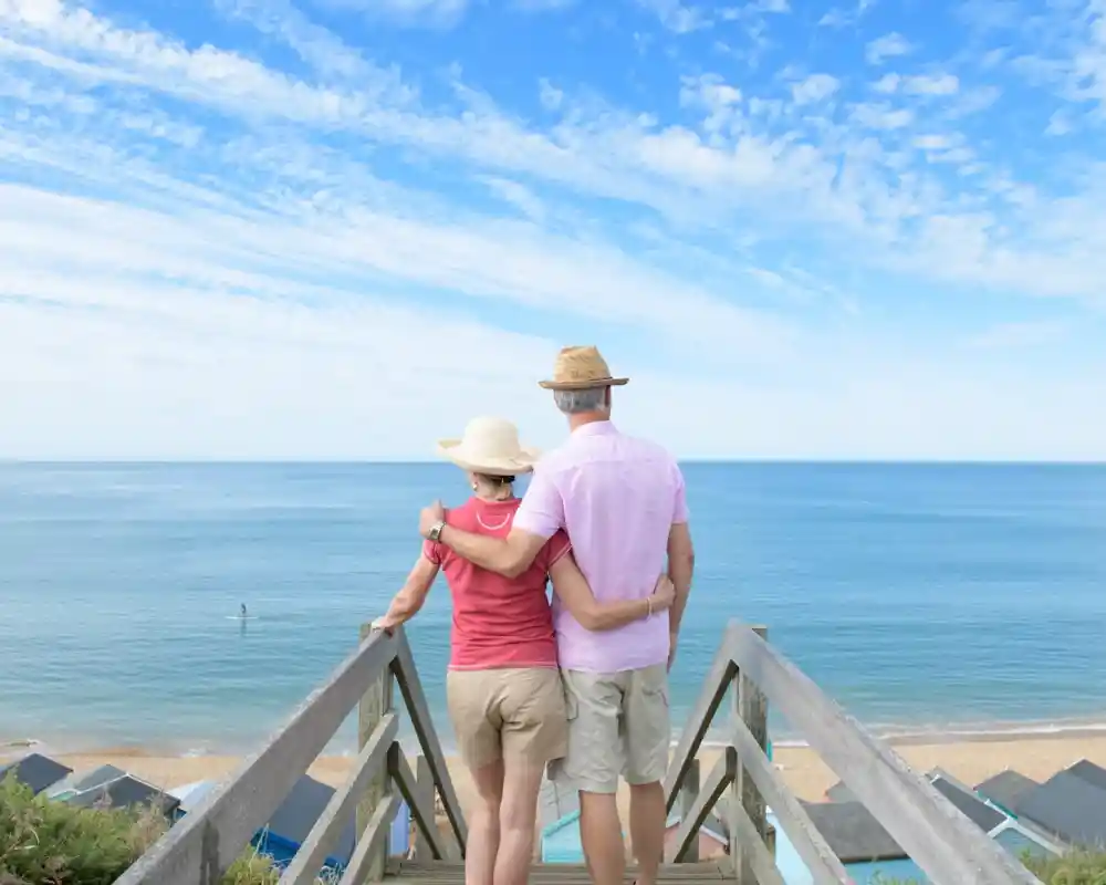 A couple stands on a wooden deck overlooking a calm sea, with the woman in a hat and the man in a light shirt. They embrace while gazing at the horizon under a blue sky with scattered clouds.