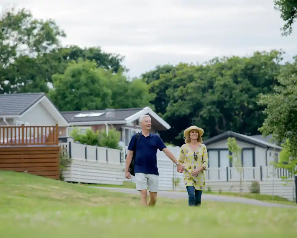 An older couple walking hand in hand along a grassy path, surrounded by trees and modern homes. The man wears a blue shirt and shorts, while the woman wears a yellow patterned dress and a wide-brimmed hat.