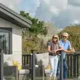 A cheerful elderly couple stands on a balcony, smiling and enjoying the outdoors. The woman is wearing a casual outfit with a scarf, while the man is in a polo shirt. They hold drinks and appear relaxed, with a well-furnished area visible behind them and greenery in the background.