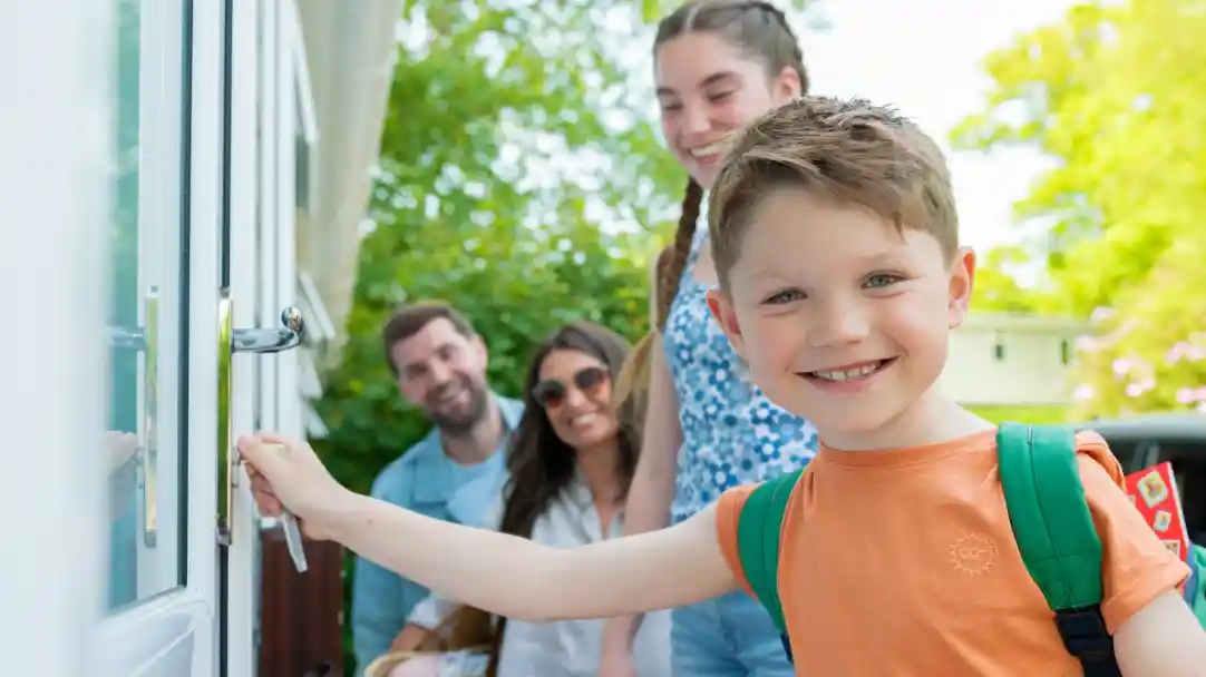 A young boy in an orange shirt and green backpack smiles as he prepares to open a door, while three adults stand behind him, enjoying the sunny outdoor setting.
