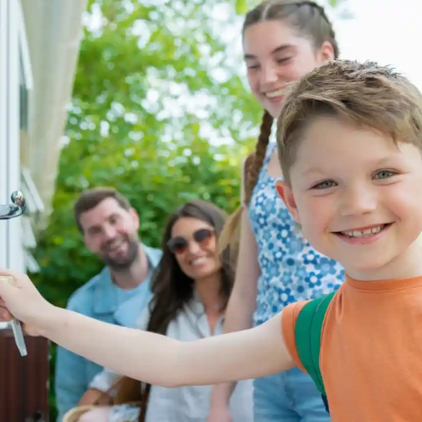 A young boy in an orange shirt and green backpack smiles as he prepares to open a door, while three adults stand behind him, enjoying the sunny outdoor setting.