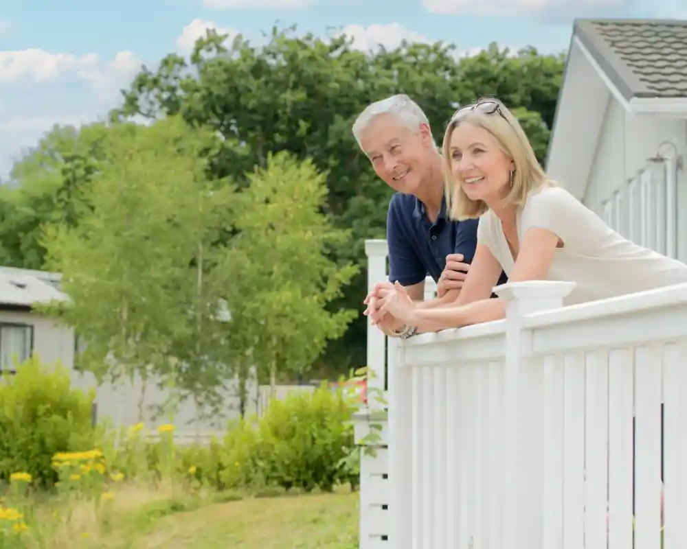 A man and a woman lean against a white fence, smiling and enjoying a sunny day in a landscaped garden. Lush greenery and flowers surround their home in the background.