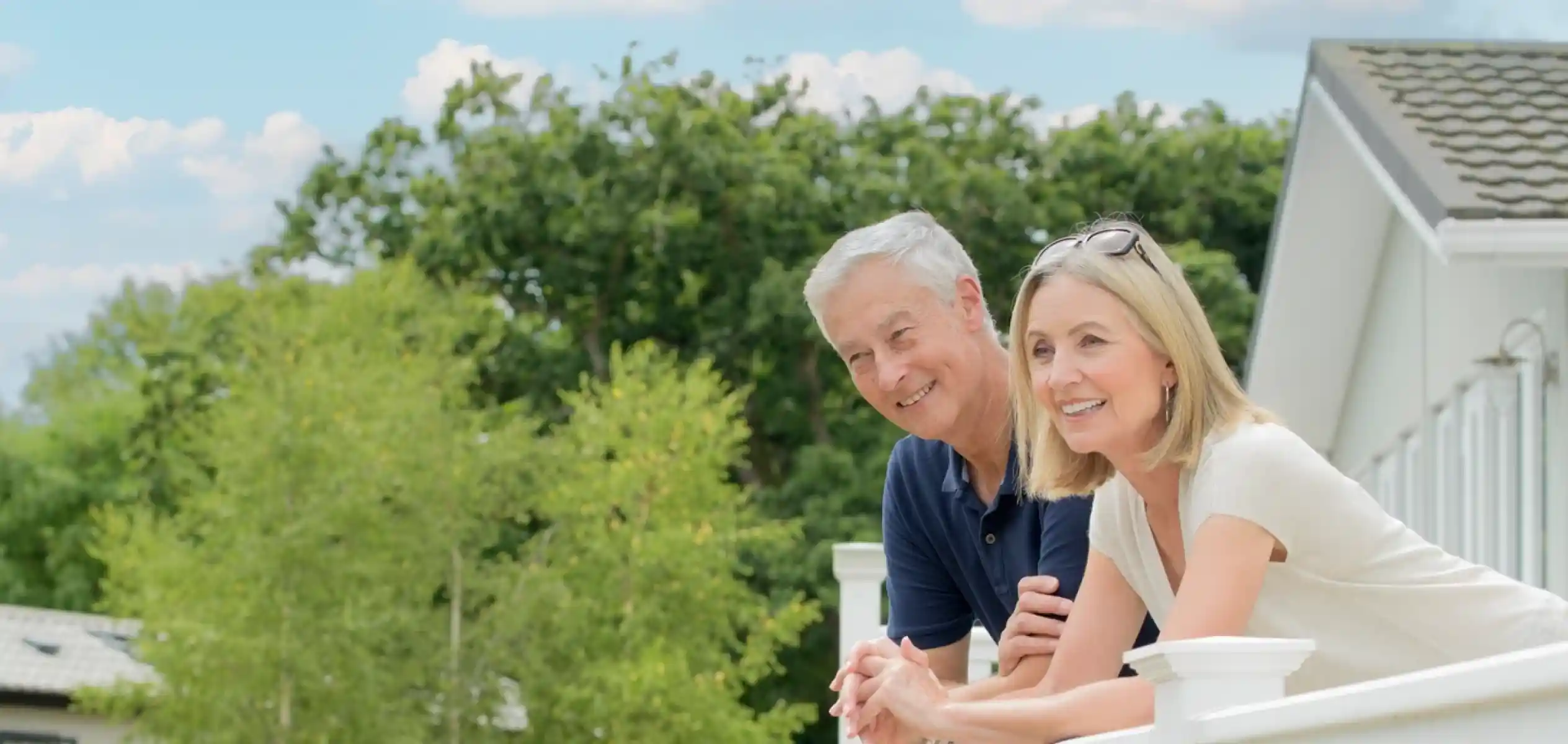 A man and a woman lean against a white fence, smiling and enjoying a sunny day in a landscaped garden. Lush greenery and flowers surround their home in the background.