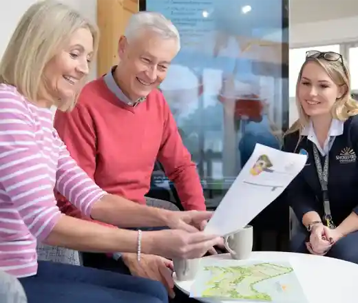 Three people are engaged in a discussion around a table, looking at documents. A woman in a pink striped sweater and a man in a red sweater smile as they examine the papers. A woman in a navy sweater with a lanyard observes and assists, all seated in a modern, well-lit setting with a digital screen in the background.