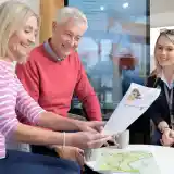Three people are engaged in a discussion around a table, looking at documents. A woman in a pink striped sweater and a man in a red sweater smile as they examine the papers. A woman in a navy sweater with a lanyard observes and assists, all seated in a modern, well-lit setting with a digital screen in the background.