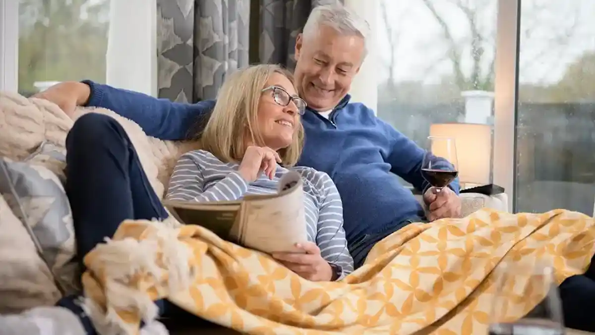 A group of five people walks together along a forest path, smiling and enjoying their time. Two adults carry a child on their back, while a black dog walks beside them. The scene is surrounded by lush greenery and autumn colors.