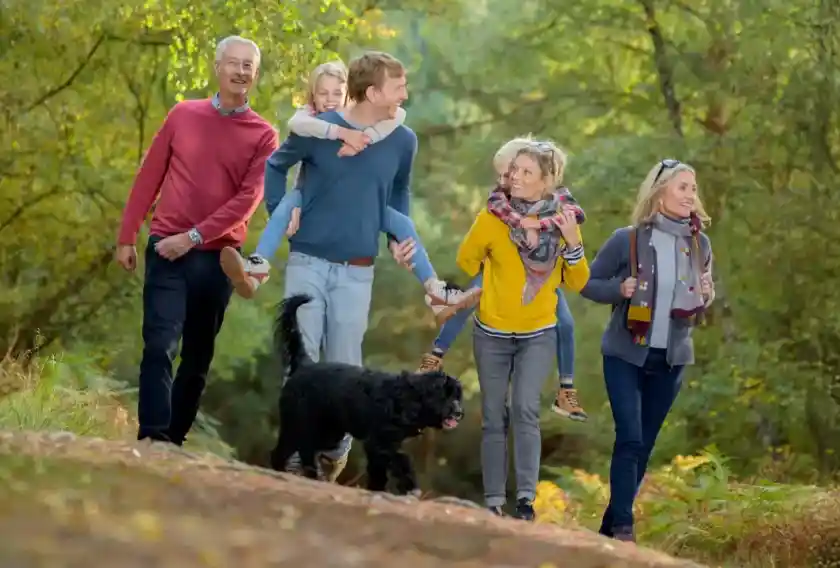 A group of five people walks together along a forest path, smiling and enjoying their time. Two adults carry a child on their back, while a black dog walks beside them. The scene is surrounded by lush greenery and autumn colors.