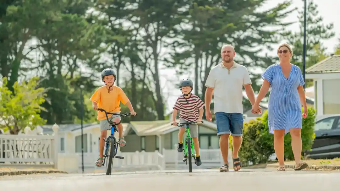 A family walks along a path in a residential area, with two young boys on bicycles. One boy wears an orange shirt and a helmet, while the other wears a striped shirt. The parents hold hands, with the father wearing a light shirt and shorts, and the mother in a blue dress. Trees and mobile homes line the background.