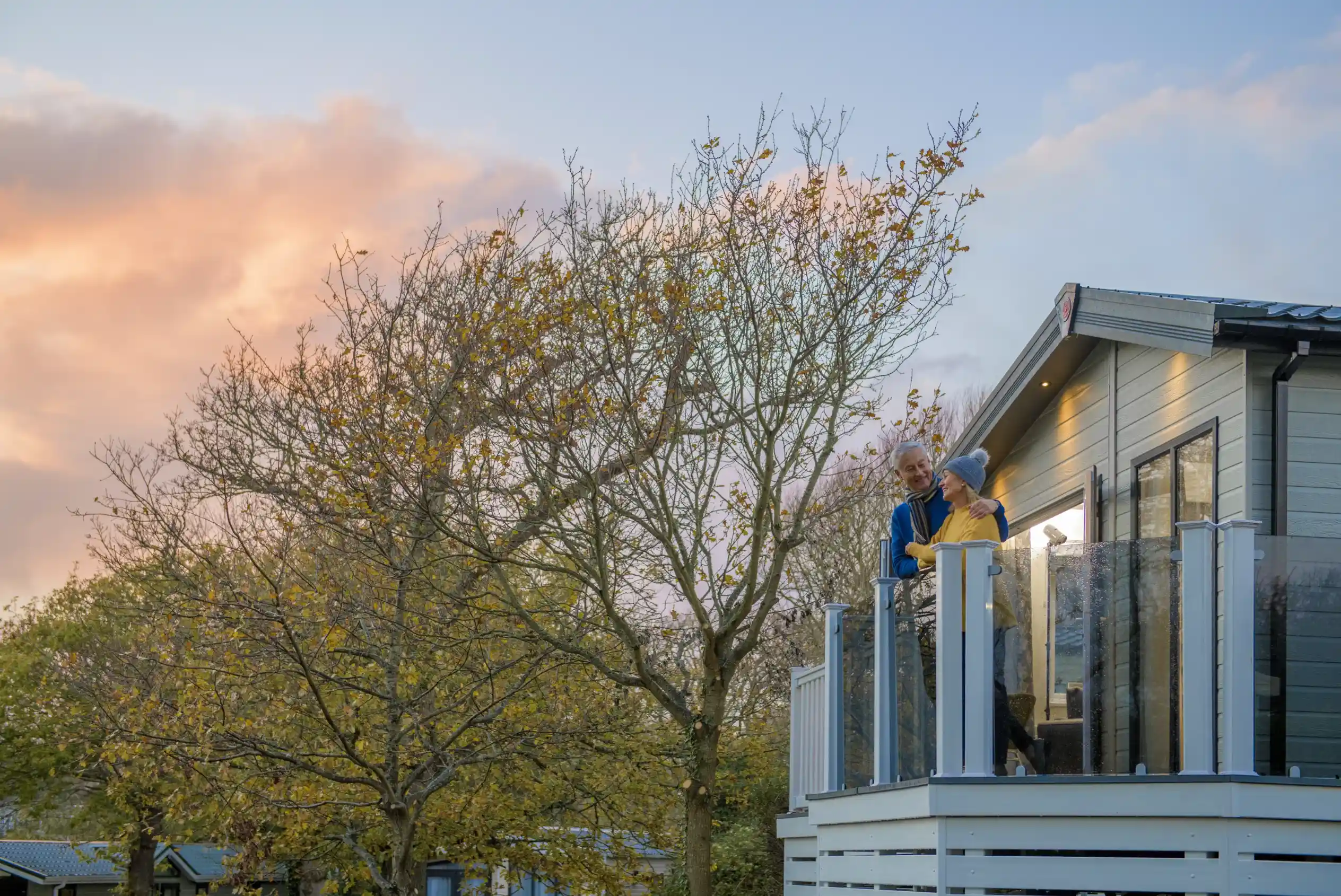 An older couple stands on a balcony, enjoying a moment together as the sun sets in the background. Trees with sparse leaves surround the house, and the sky is tinged with warm hues of orange and pink.