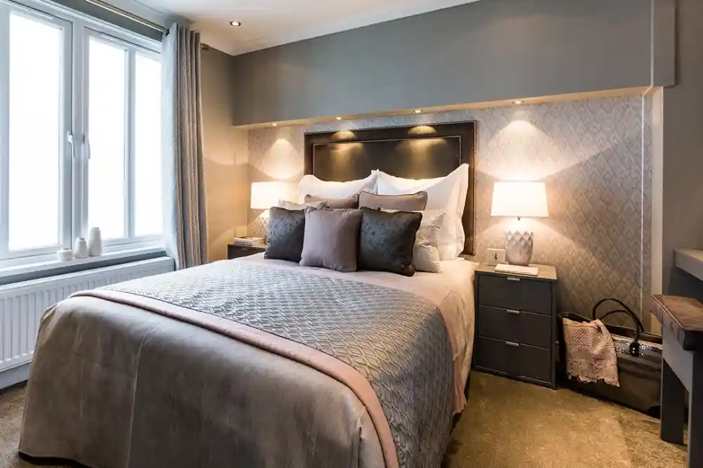 A cozy bedroom featuring a queen-sized bed with gray and blush-colored bedding, flanked by two bedside tables with lamps. Soft curtains frame a large window, and the walls have a subtle patterned texture.