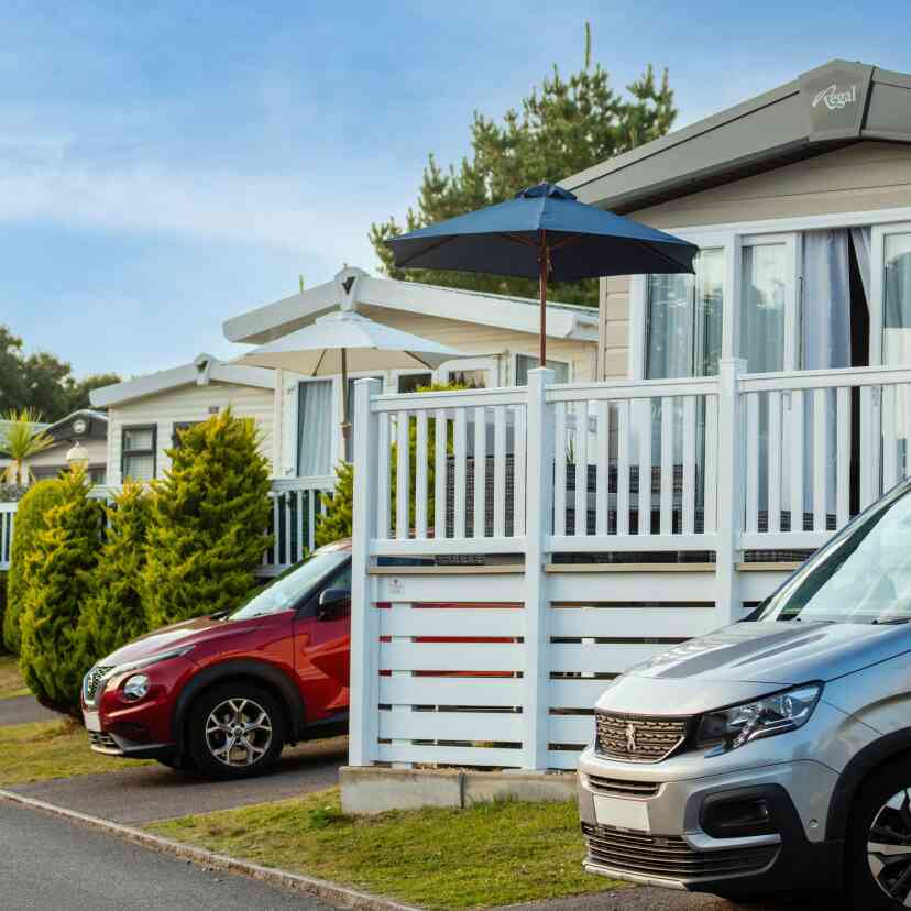 Colorful holiday caravans lined along a paved road, with an umbrella-shaded deck in front of one. A silver vehicle is parked beside a red car, with manicured hedges lining the pathway. Bright blue sky overhead.