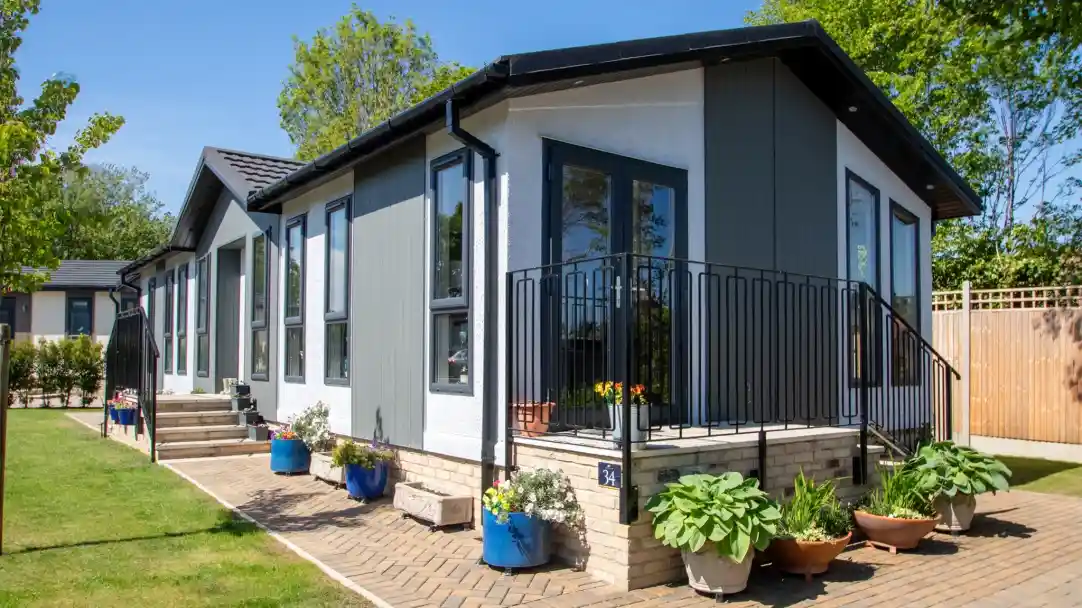 Modern house with a stylish design, featuring a porch with a black railing. Colorful potted plants decorate the entrance, and a well-kept lawn surrounds the property. Bright blue sky and greenery in the background.