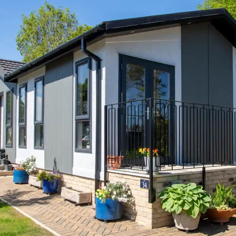 Modern house with a stylish design, featuring a porch with a black railing. Colorful potted plants decorate the entrance, and a well-kept lawn surrounds the property. Bright blue sky and greenery in the background.