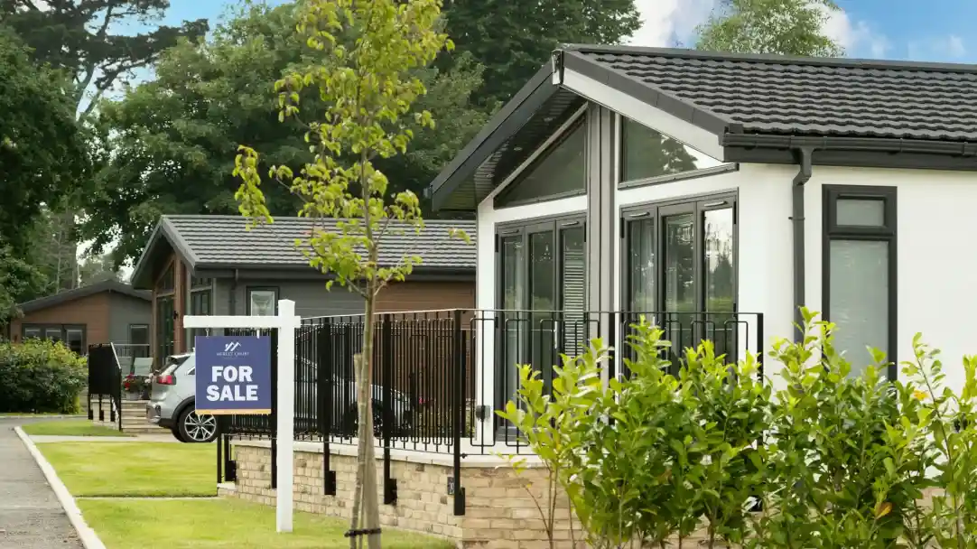 A modern house with large windows and a black roof is shown. In front, a "For Sale" sign stands beside a landscaped area with small bushes. Nearby, several similar houses are visible along a well-maintained pathway.