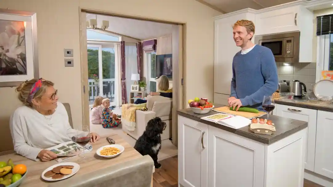 A man stands at a kitchen island, smiling while preparing food. A woman sits at the dining table with a glass of wine and a magazine, looking at him. In the background, children are playing on the floor, and another adult sits on a couch. A dog is near the kitchen island. The space is bright and cozy, with decorative elements visible.