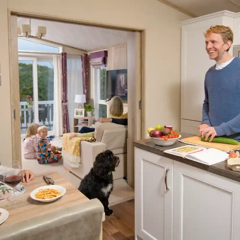 A man stands at a kitchen island, smiling while preparing food. A woman sits at the dining table with a glass of wine and a magazine, looking at him. In the background, children are playing on the floor, and another adult sits on a couch. A dog is near the kitchen island. The space is bright and cozy, with decorative elements visible.