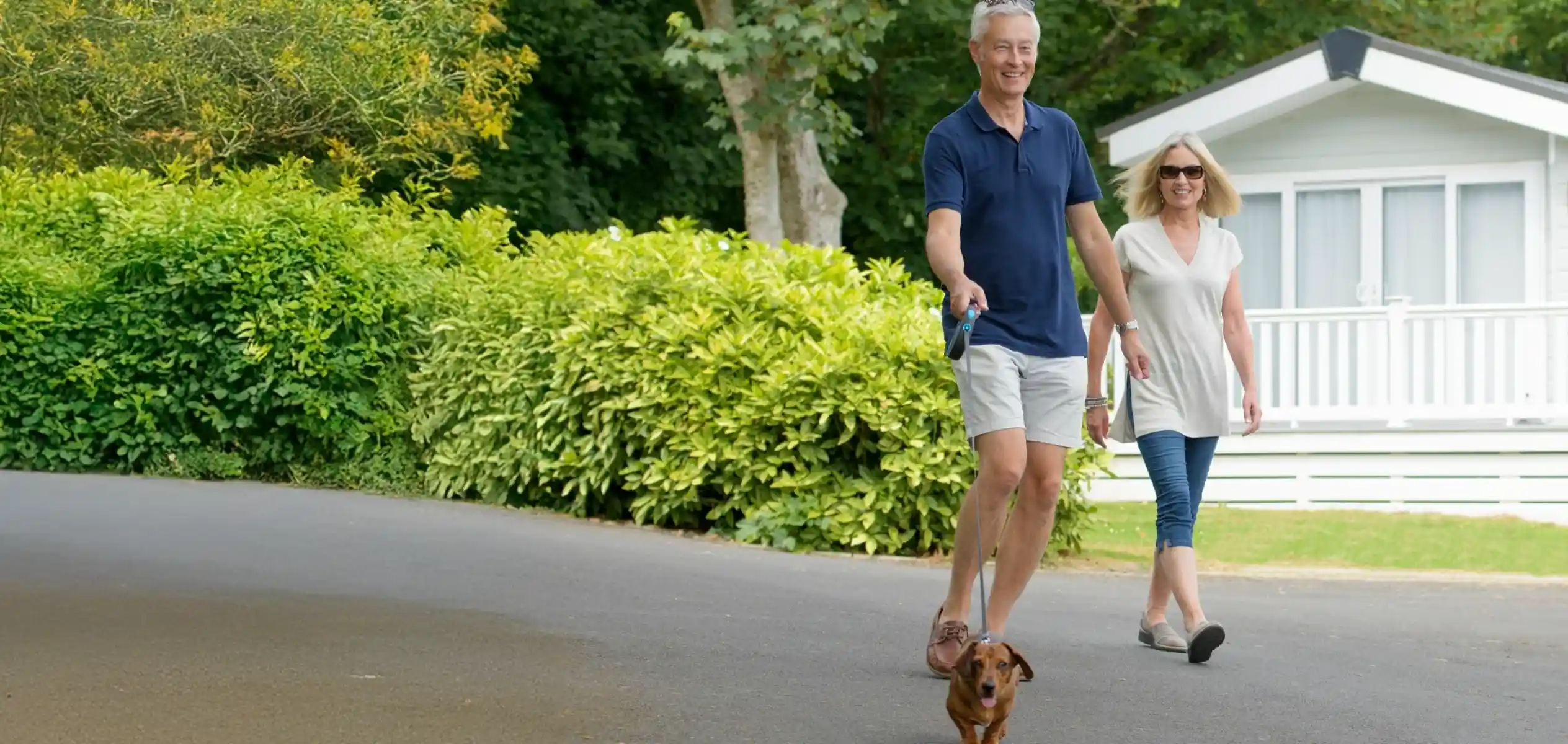 A smiling couple walks a small dog along a scenic path surrounded by lush greenery and a white static holiday home in the background. The man wears a blue polo shirt and shorts, while the woman is dressed in a light top and jeans.