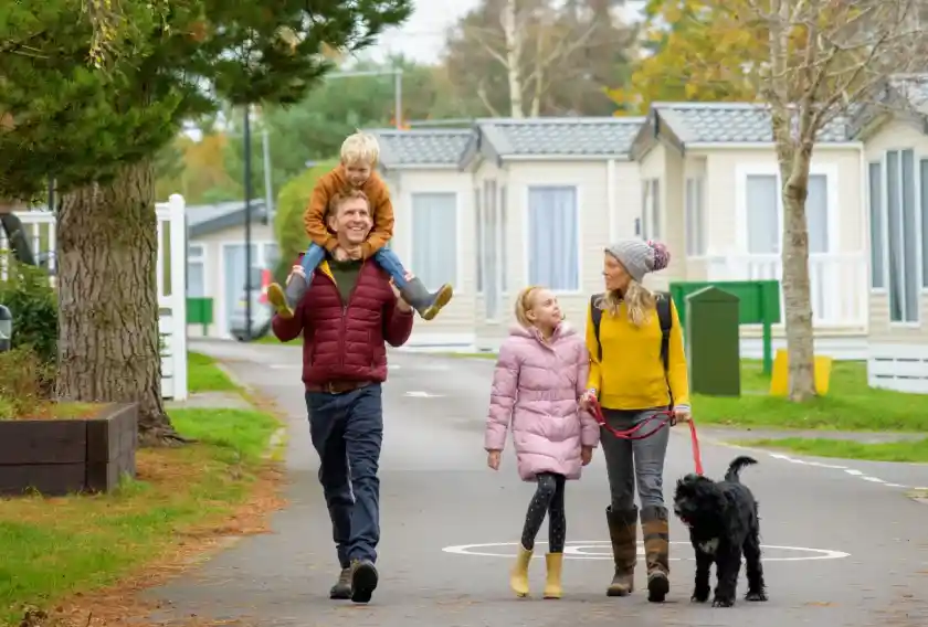A family walks down a path in a holiday park. A man carries a young boy on his shoulders, while a girl and a woman walk alongside with a dog. The environment features caravan homes and greenery, creating a relaxed atmosphere.
