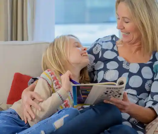A woman and a young girl sit on a couch, both smiling and enjoying a moment together. The girl leans against the woman's shoulder, holding a book, while the woman laughs, creating a warm and joyful atmosphere.
