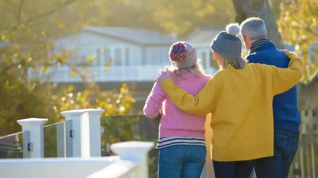A group of three people stands together on a sunny balcony, looking out at a scenic view. The person on the left wears a pink sweater and a colorful knit hat, while the person in the middle is dressed in a bright yellow sweater. The individual on the right wears a gray beanie and a blue sweater. Warm sunlight filters through the trees, casting a golden glow.
