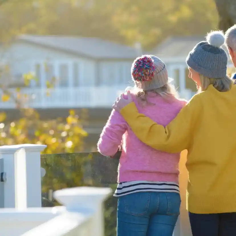 A group of three people stands together on a sunny balcony, looking out at a scenic view. The person on the left wears a pink sweater and a colorful knit hat, while the person in the middle is dressed in a bright yellow sweater. The individual on the right wears a gray beanie and a blue sweater. Warm sunlight filters through the trees, casting a golden glow.
