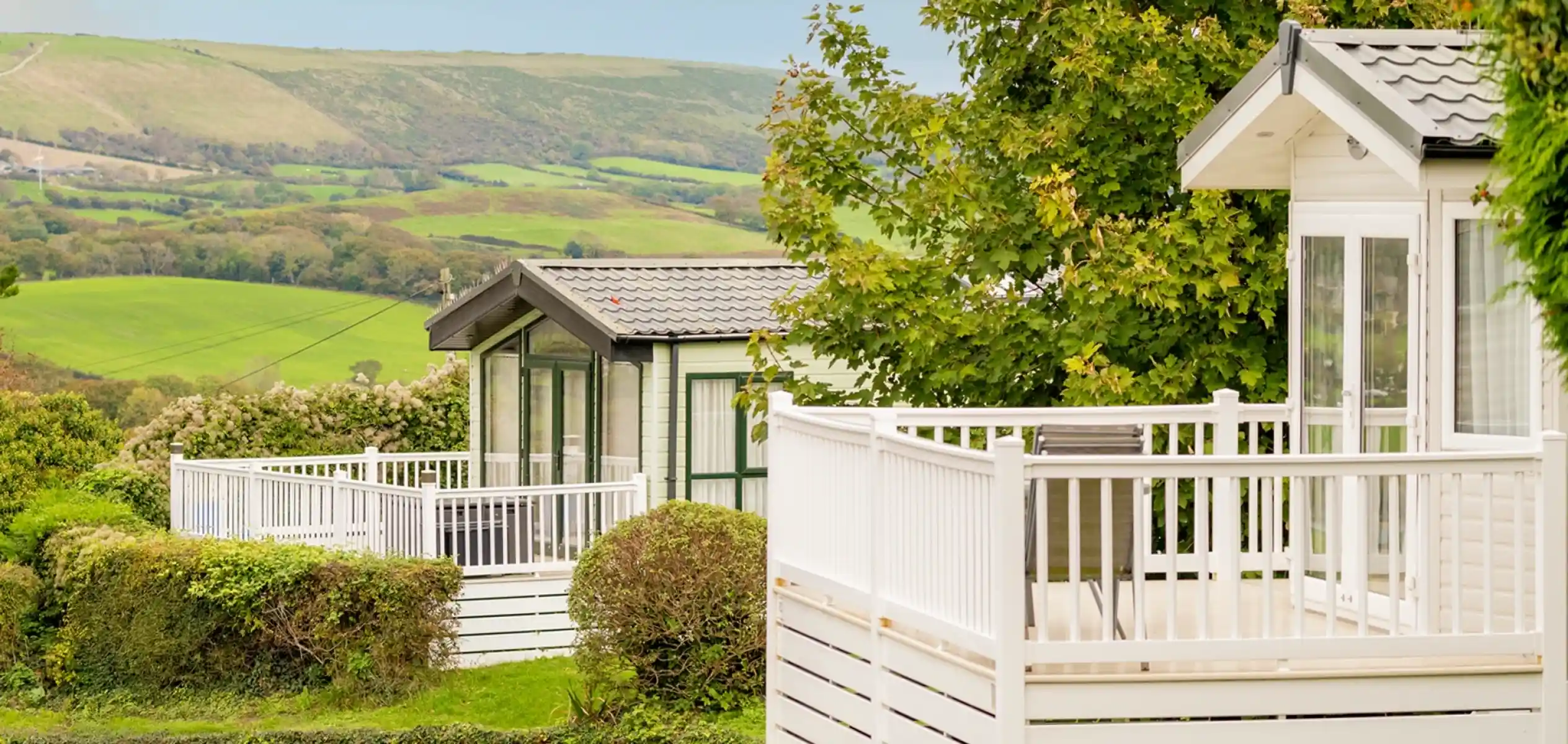 Two modern static caravans with white railings nestled in a lush green landscape. Rolling hills and trees surround the area, creating a tranquil atmosphere.