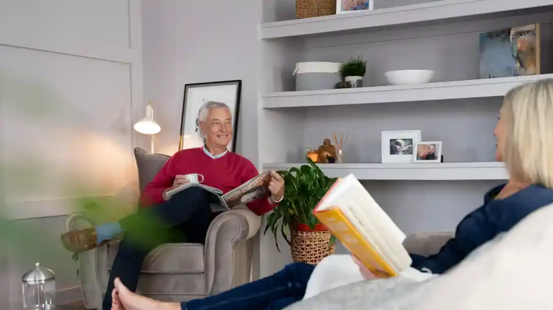An older man sits comfortably in an armchair, smiling and holding a book, while a middle-aged woman relaxes on a nearby sofa, also holding a book. The room features light-colored walls, shelves with decorative items, and green plants.