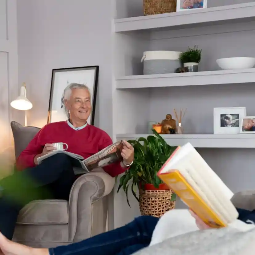 An older man sits comfortably in an armchair, smiling and holding a book, while a middle-aged woman relaxes on a nearby sofa, also holding a book. The room features light-colored walls, shelves with decorative items, and green plants.