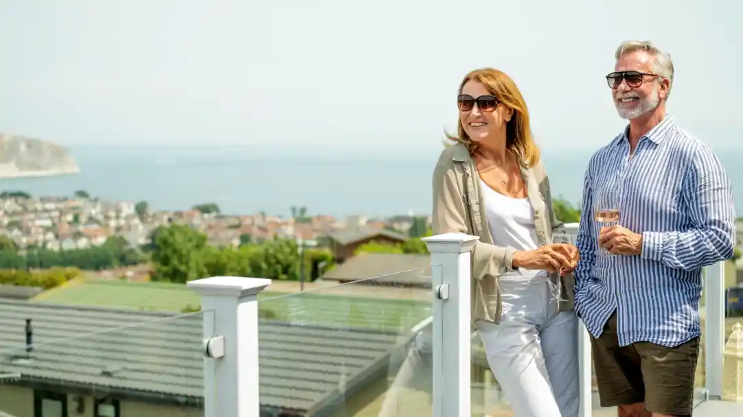 A smiling woman and man stand together on a balcony with a scenic view of the coast and town below. The woman is wearing sunglasses and a light outfit, while the man is dressed in a striped shirt and shorts, holding a glass. They appear relaxed and enjoying each other's company.