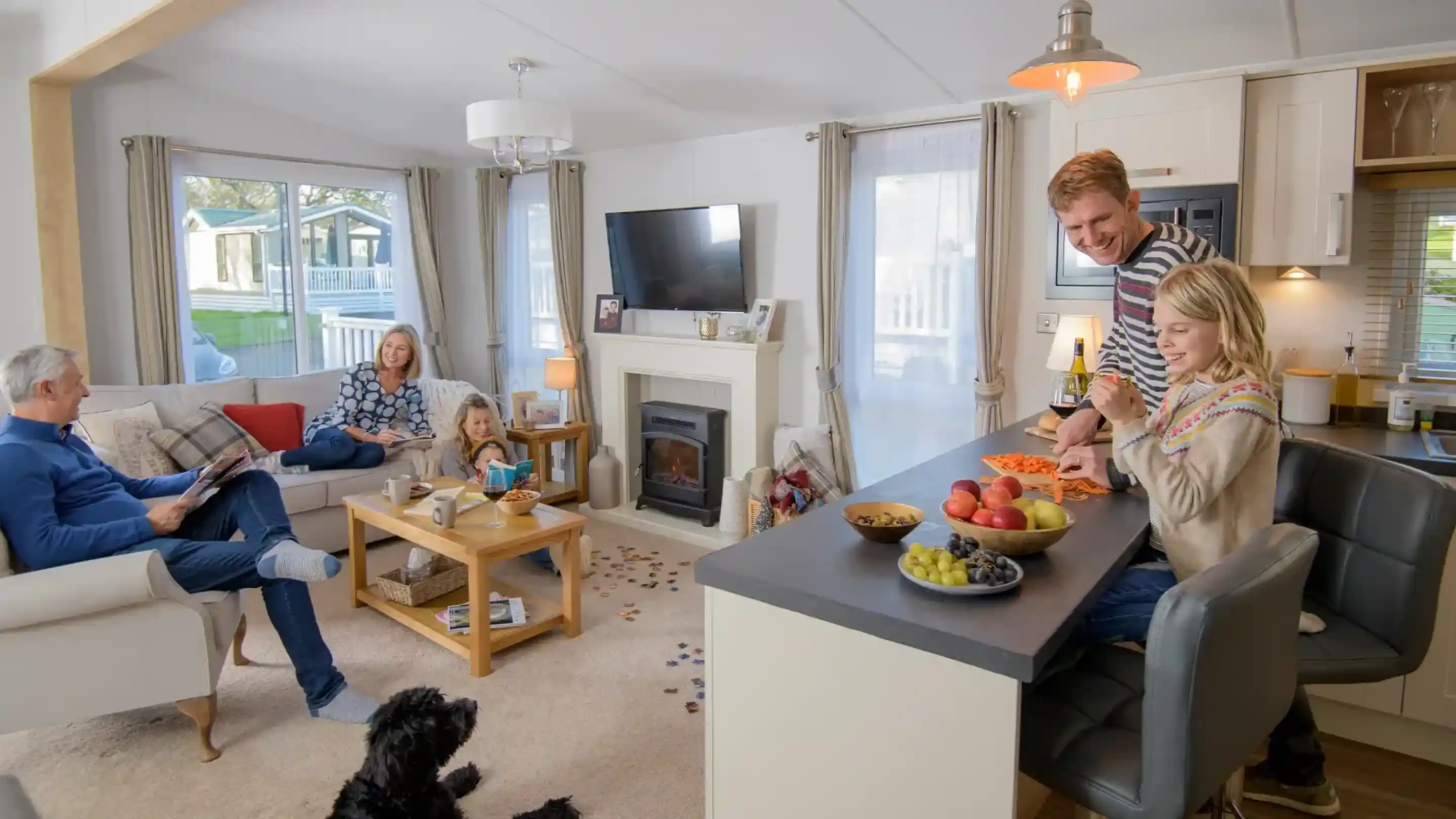A family enjoying time together in a cosy living room. A man prepares food at a kitchen island while a girl helps with fruits. A woman and another girl are seated on a sofa, and a man reads a magazine nearby. A black dog sits on the floor.