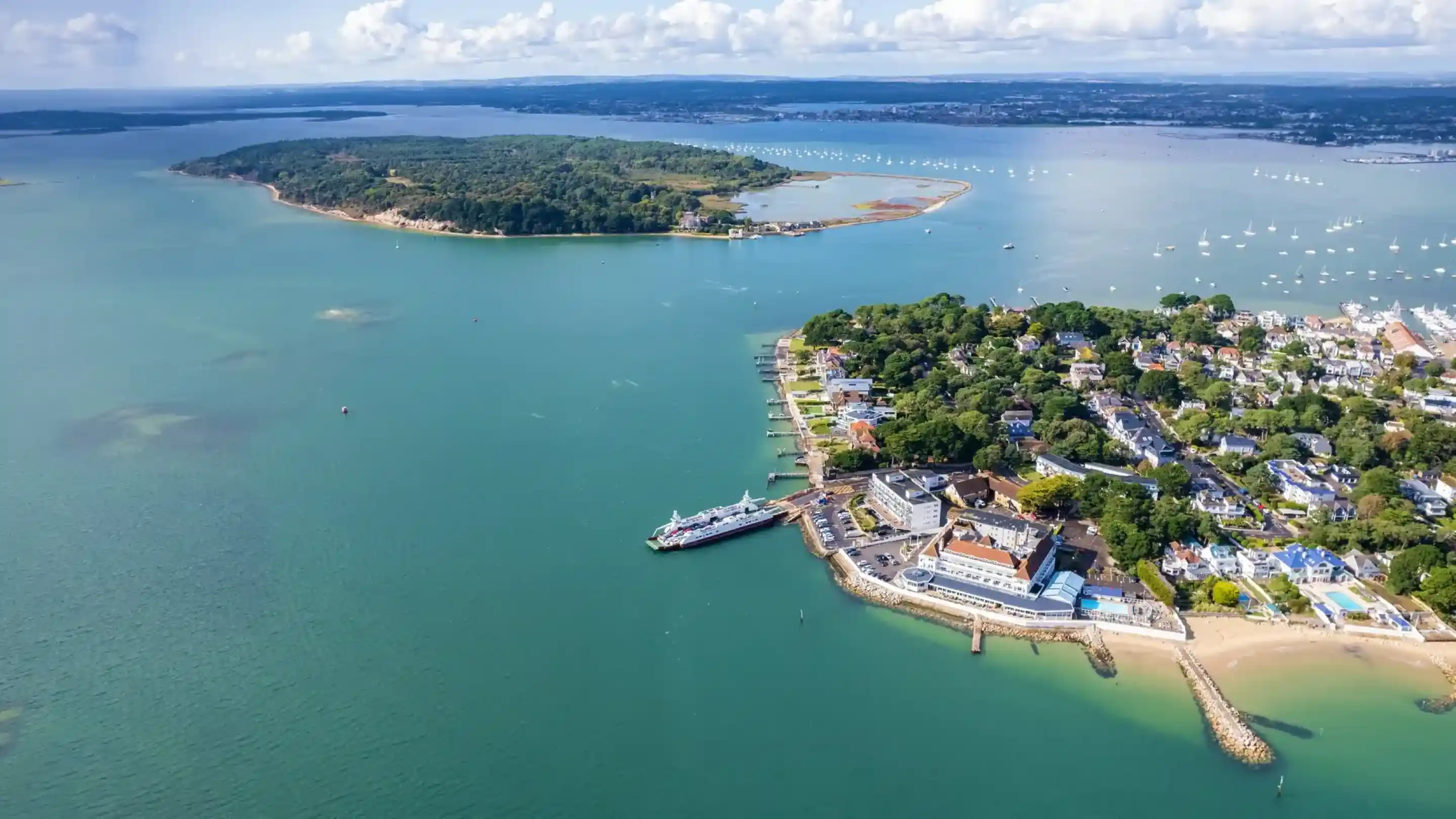 Aerial view of Poole Harbour featuring a peninsula with buildings and a marina. Clear blue waters surround the land, with boats scattered throughout. In the background, lush green hills rise above the water, and fluffy white clouds dot the sky.