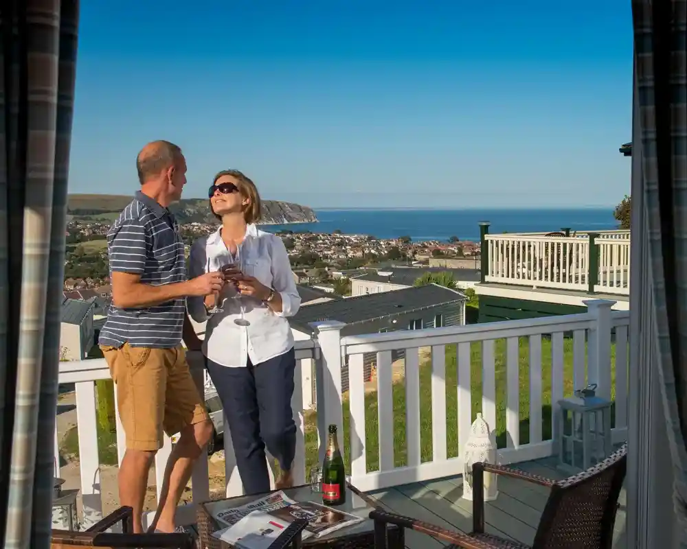 A couple enjoys drinks on a balcony with a view of the ocean and coastline. The woman is smiling and looking at the man, who is casually dressed. A bottle of champagne and glasses are on a nearby table. Clear blue skies enhance the serene atmosphere.