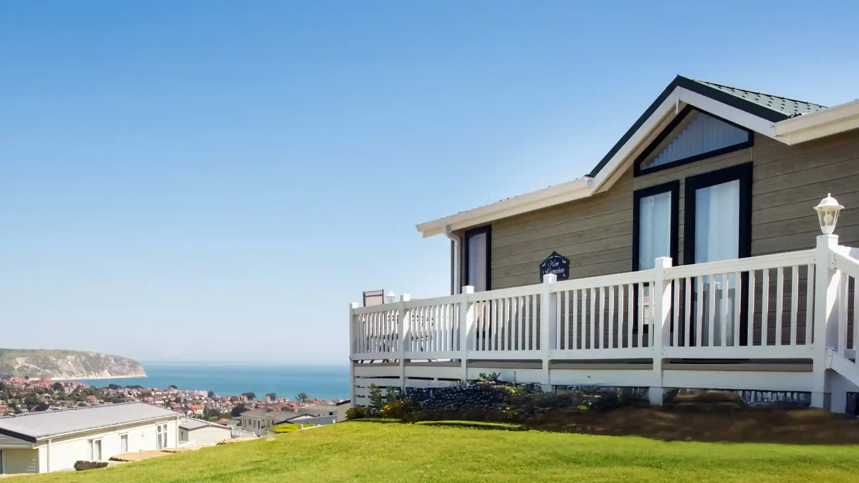 A modern house with a wooden exterior and a white railing, overlooking a scenic coastline and blue ocean under a clear sky. Lush green grass surrounds the house.