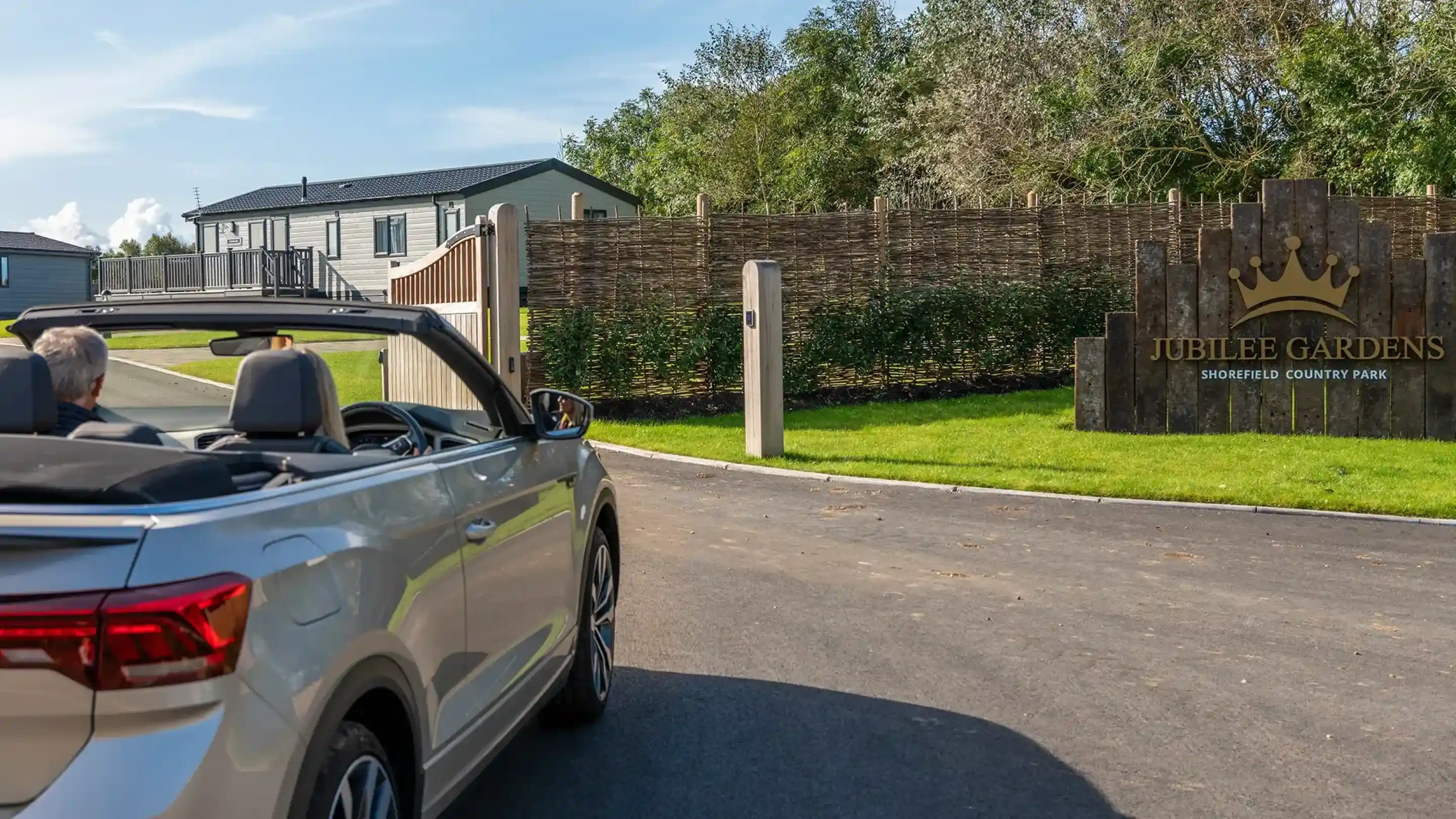 A silver convertible car approaches the entrance of Jubilee Gardens, a country park. A wooden sign with a crown logo marks the entrance, surrounded by greenery and a paved road leading in.