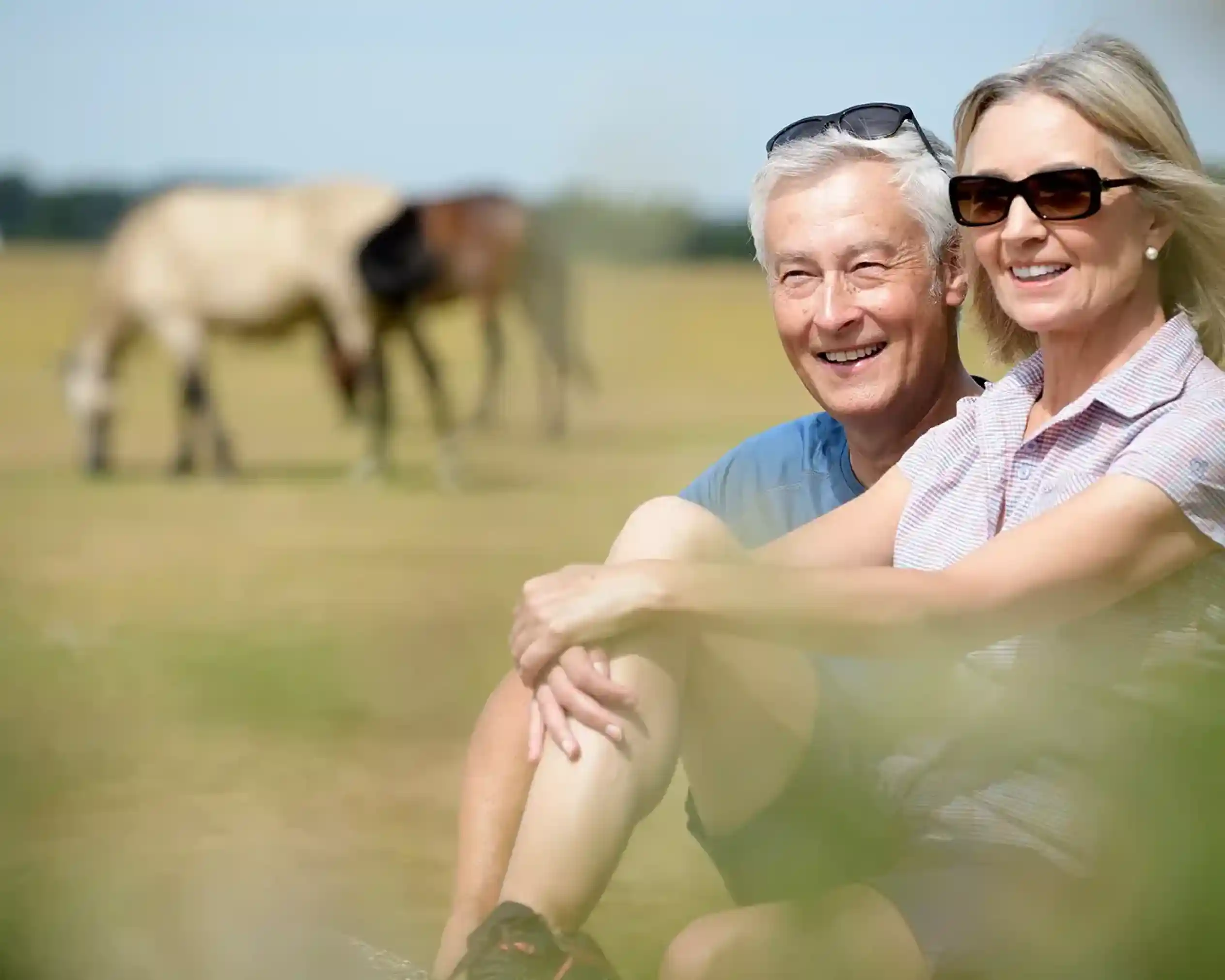 An older couple smiles while sitting together outdoors. They are casually dressed, with the woman wearing sunglasses. In the background, horses graze in a grassy field.