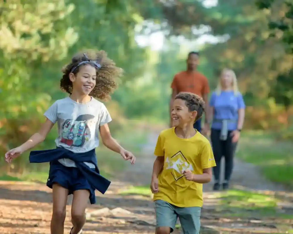 Two children are joyfully running along a forest path. The girl has curly hair and wears a gray T-shirt and a blue skirt, while the boy, with short curly hair, is dressed in a yellow T-shirt and light shorts. In the background, two adults are walking, one wearing an orange shirt and the other in a purple shirt. The scene is bright and vibrant, surrounded by lush green trees.