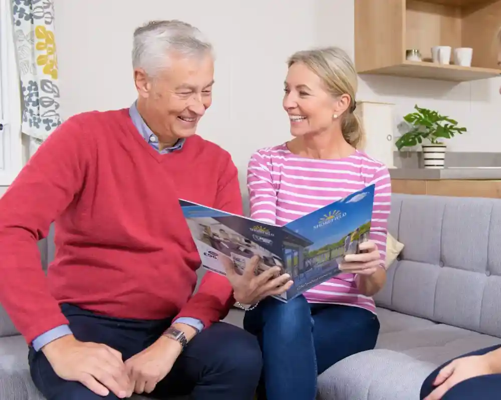 A man and a woman sit on a grey couch looking at a brochure, smiling and engaged in conversation. A woman in a dark sweater sits across from them, offering assistance. The setting is a bright, cosy living space with simple décor.