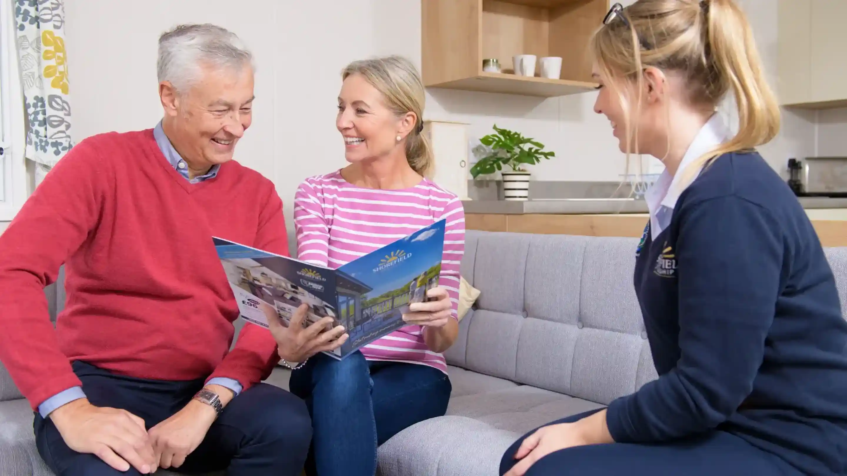 A man and a woman sit on a grey couch looking at a brochure, smiling and engaged in conversation. A woman in a dark sweater sits across from them, offering assistance. The setting is a bright, cosy living space with simple décor.