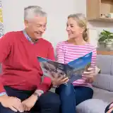 A man and a woman sit on a grey couch looking at a brochure, smiling and engaged in conversation. A woman in a dark sweater sits across from them, offering assistance. The setting is a bright, cosy living space with simple décor.